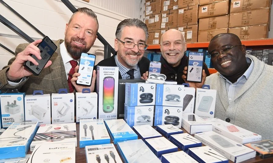 Four men smiling and displaying electronic gadgets and accessories on a table and holding their phones, in a warehouse setting.