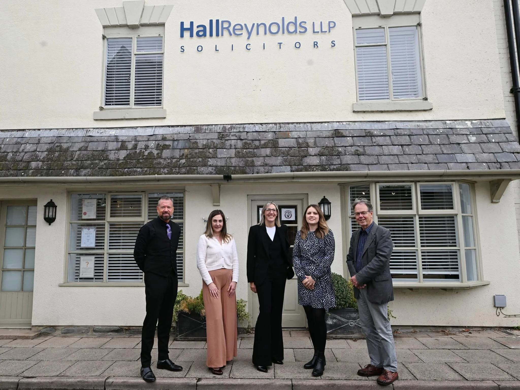 Group of five professionals standing outside in front of a building with a sign that reads 'Hall Reynolds LLP Solicitors'.
