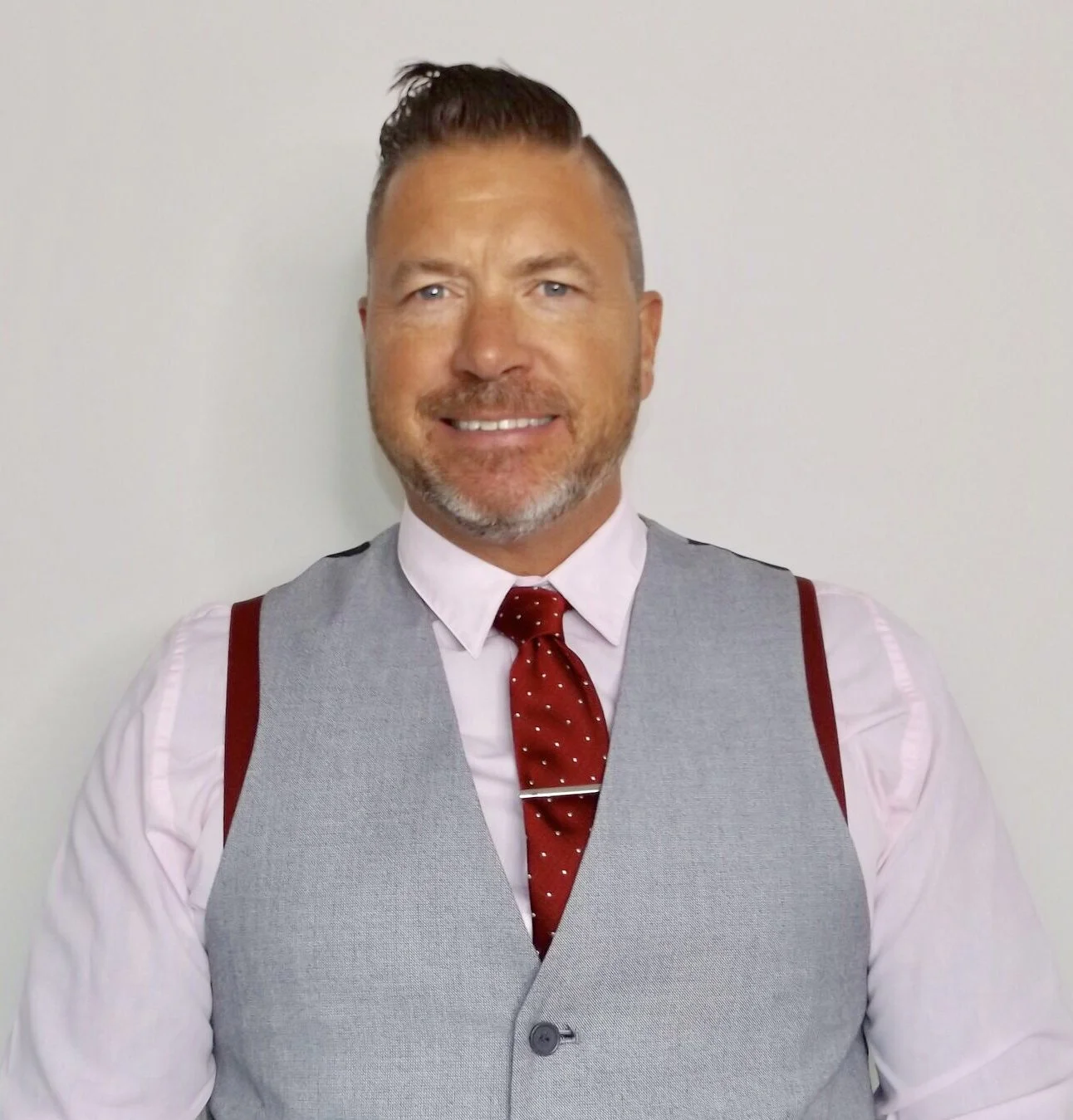 A man with light skin, blue eyes, and a beard, smiling, wearing a pink dress shirt, a gray vest, a red tie with white polka dots, and suspenders, standing against a plain white wall.