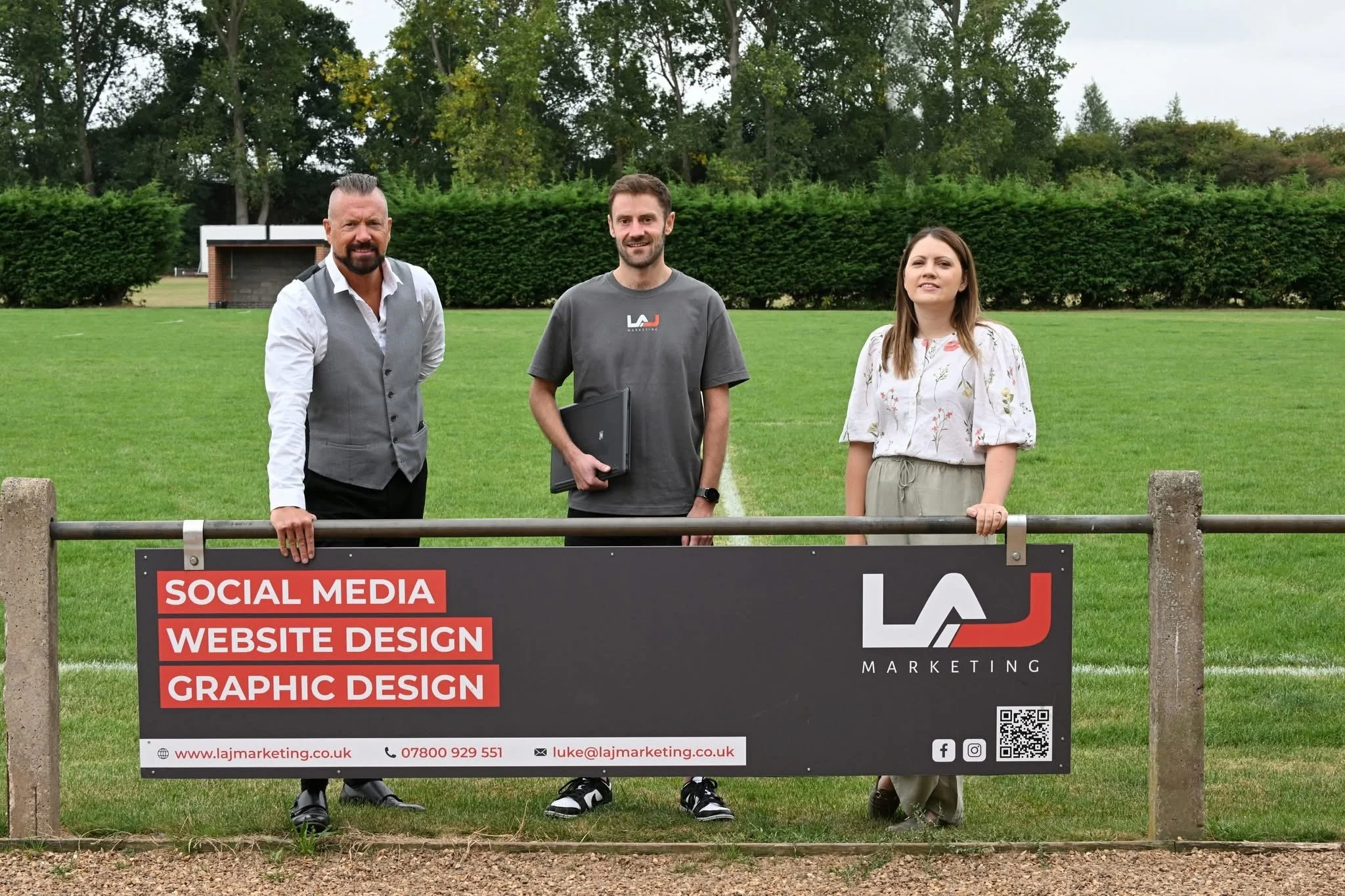 Three people standing behind a sign on a grassy field with trees in the background. The sign advertises social media, website design, and graphic design services from LAJ Marketing, with contact information including a website, phone number, email, and social media icons.