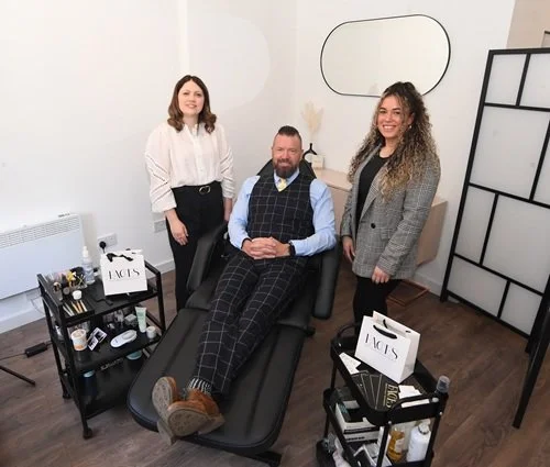 A man sitting on a treatment bed with two women standing beside him in a modern room, with beauty products on tables nearby.