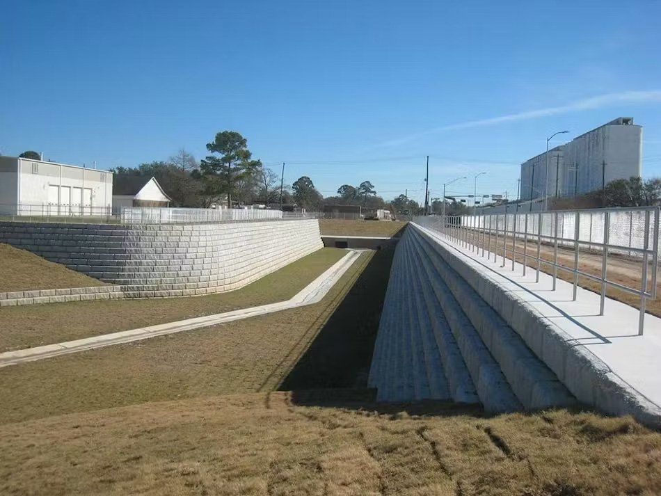 Concrete drainage system with sloped walls and a walkway, surrounded by grass and industrial buildings, under a clear blue sky.