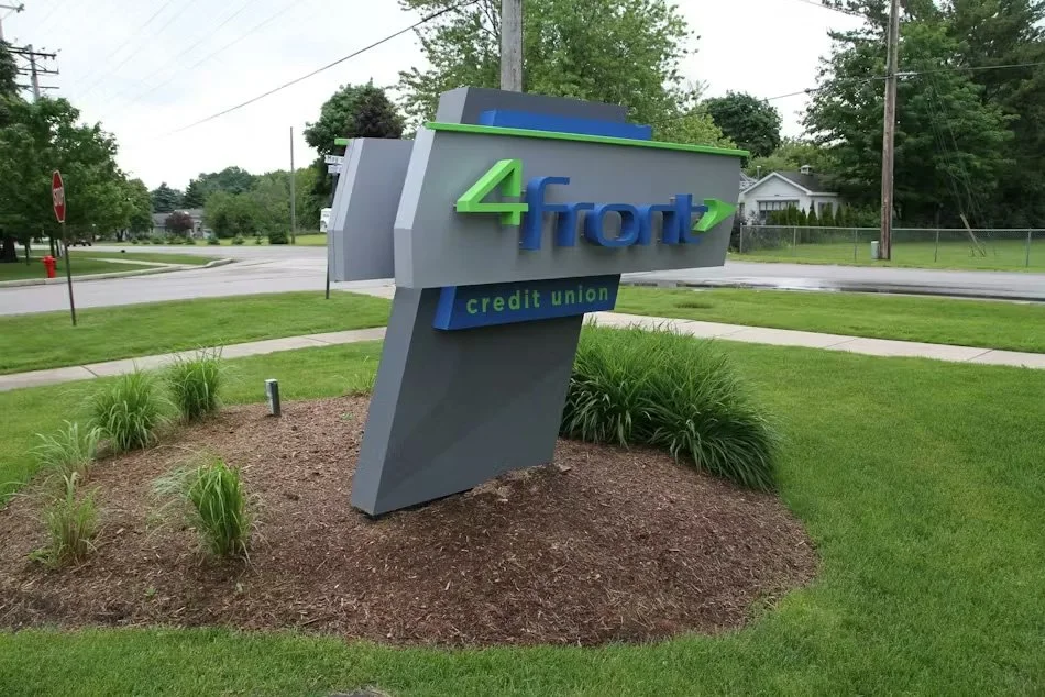 A modern outdoor sign for Frost Credit Union with green and blue lettering, set on a landscaped grassy area with small plants, with a street, trees, and houses in the background.