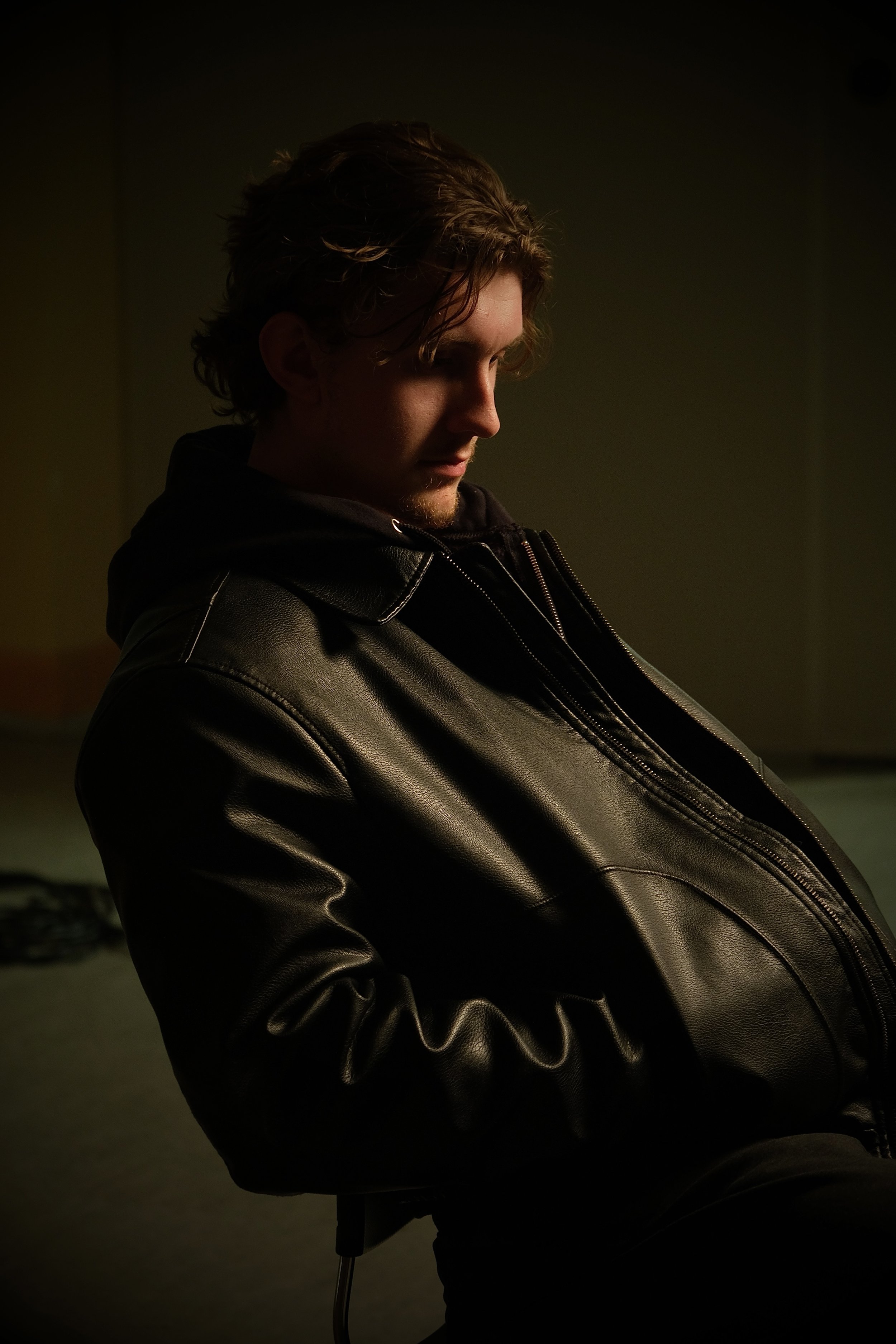 A young man with wavy brown hair, wearing a black leather jacket, sitting in a dimly lit room with a dark background, looking downward.