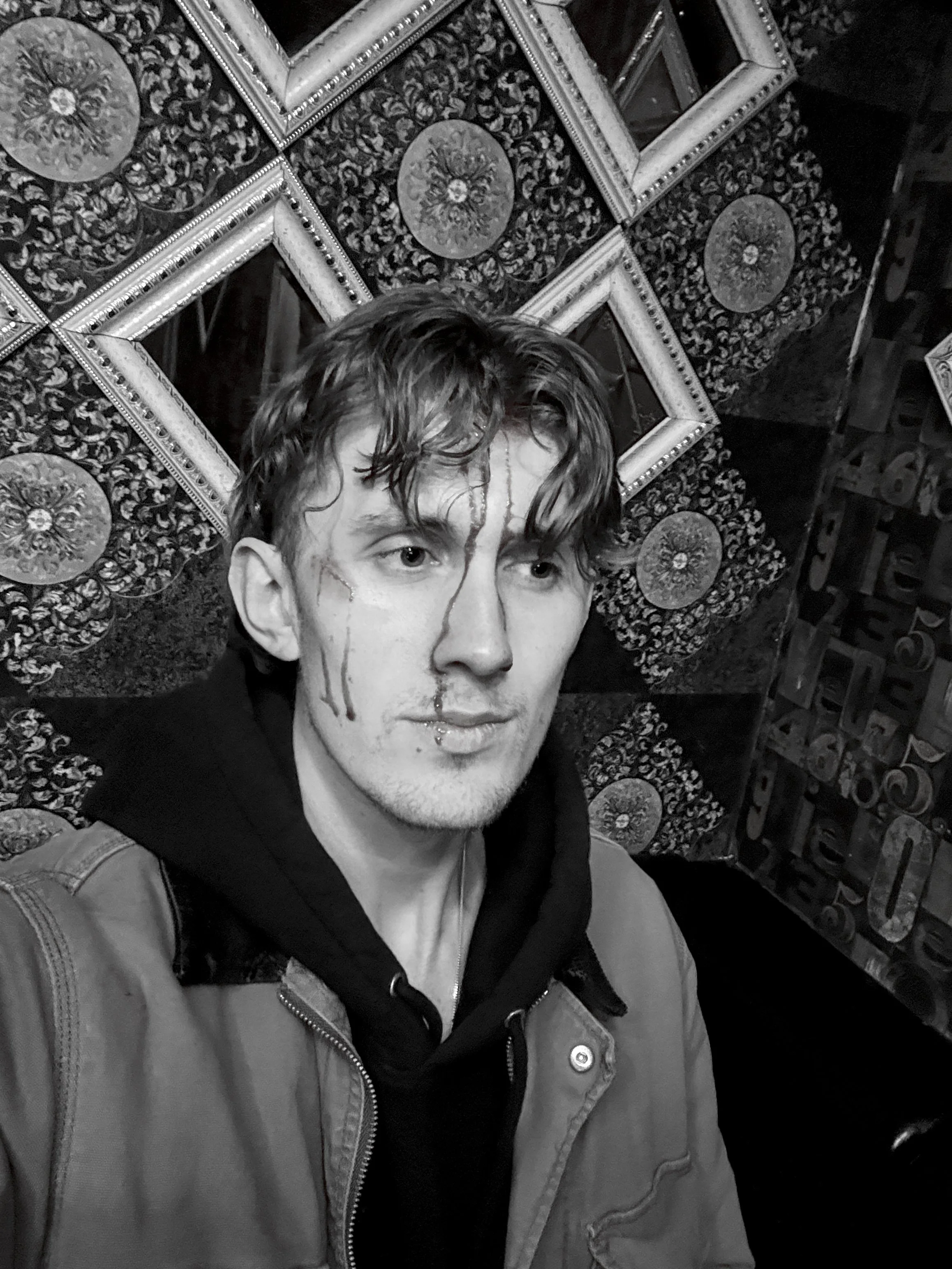 Black and white photo of a young man with wavy hair, slight facial hair, and a serious expression. He is wearing a hoodie and a jacket. Behind him, there is an ornate wall decoration with intricate patterns, framed mirrors, and decorative tiles.