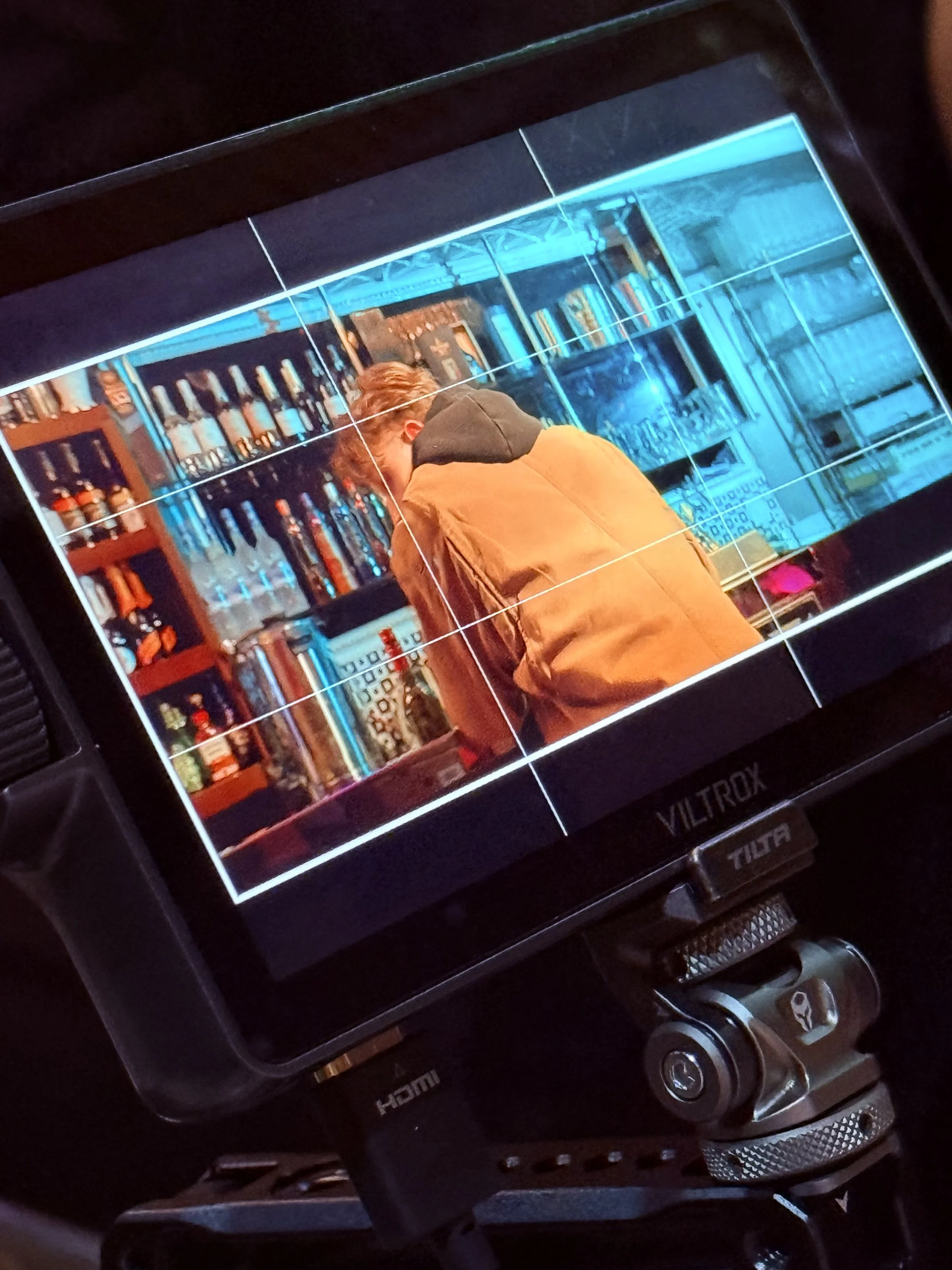 A camera viewfinder showing a person with brown hair wearing a tan jacket and black hoodie sitting at a bar with liquor bottles on shelves behind them.