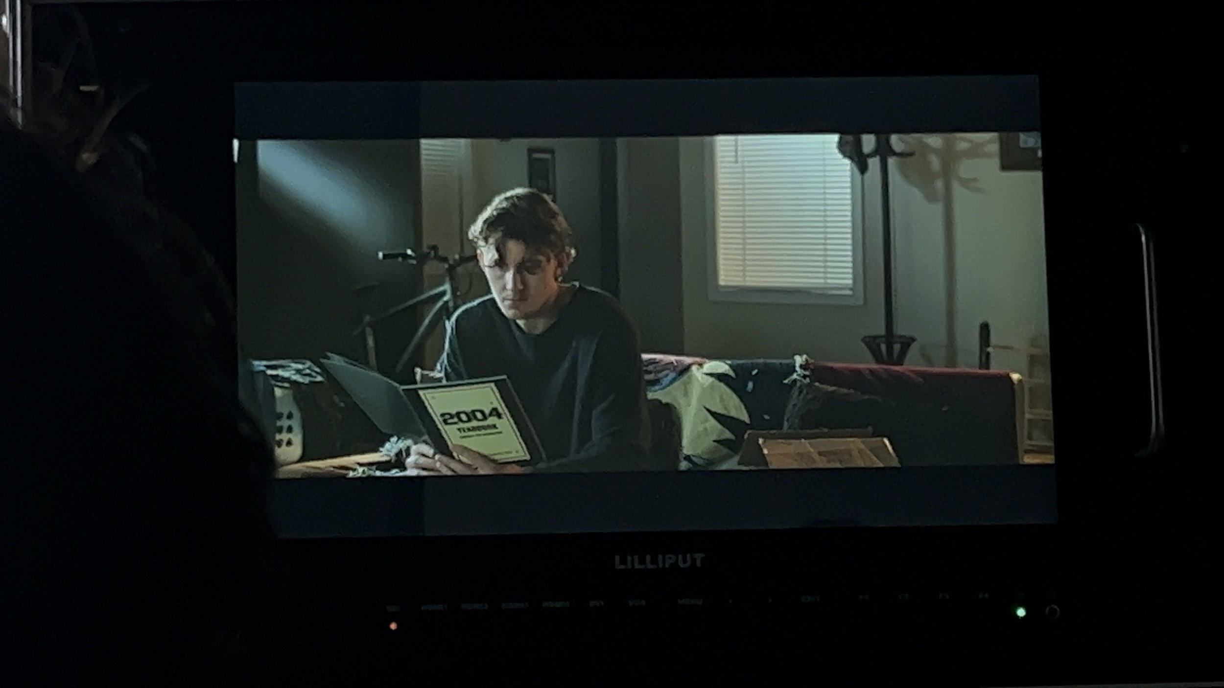 Young man with curly hair reading a booklet labeled '2004' in a dimly lit room with a bicycle and window in the background.