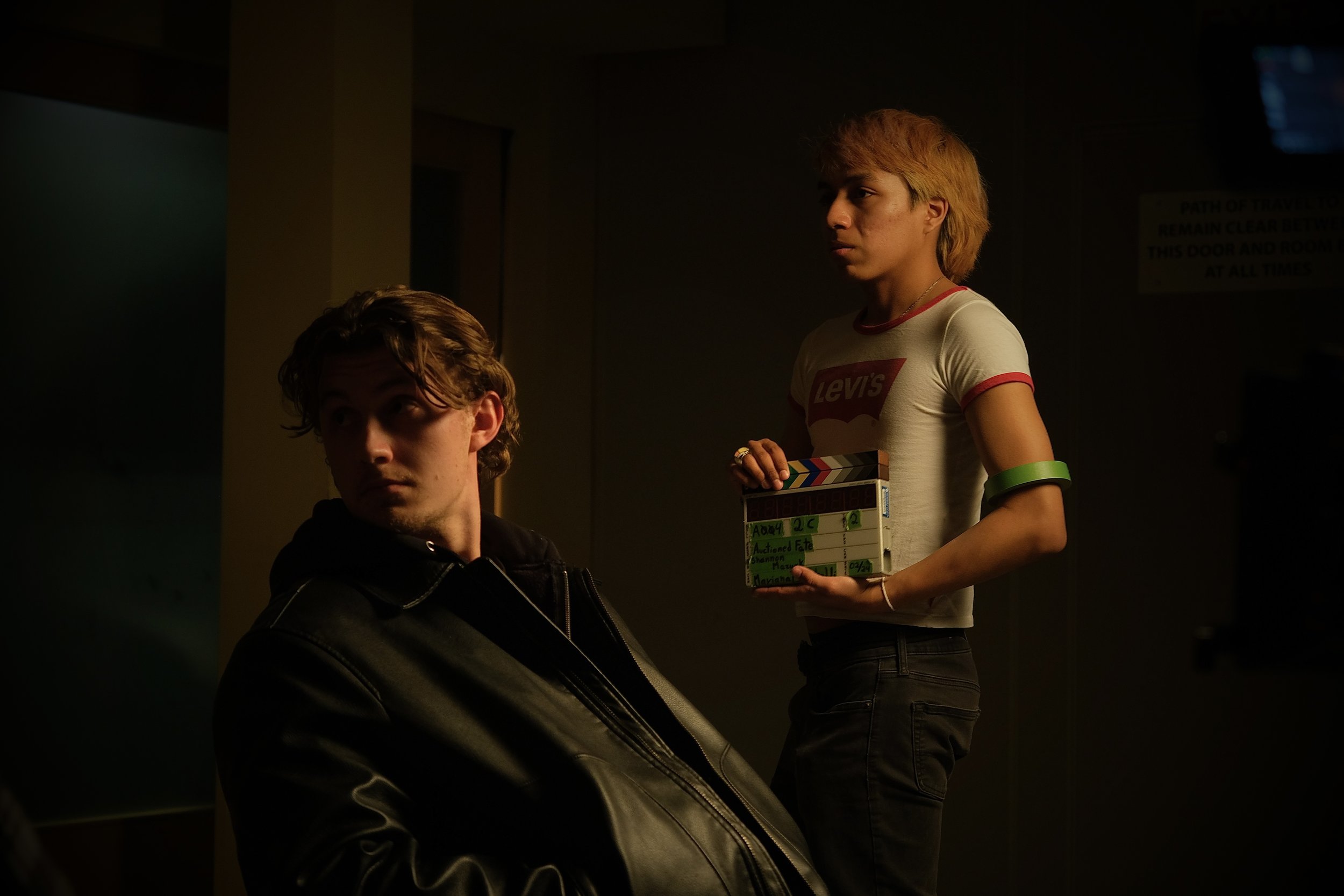 Two people on a film set, one seated in a leather jacket in shadow and one standing with a striped shirt holding a clapperboard in a dimly lit room.
