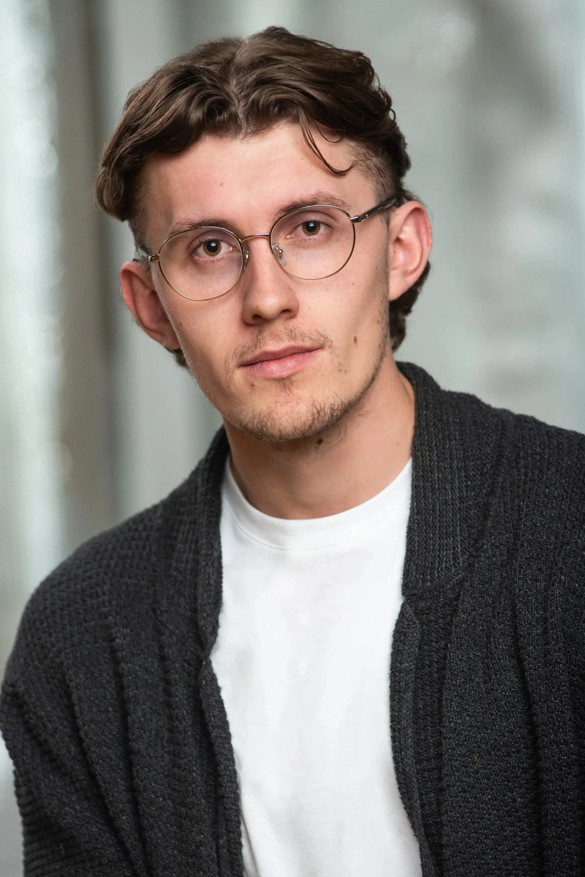 A young man with glasses, brown hair, and a light complexion wearing a white T-shirt and a dark textured cardigan, standing indoors with a blurred background.