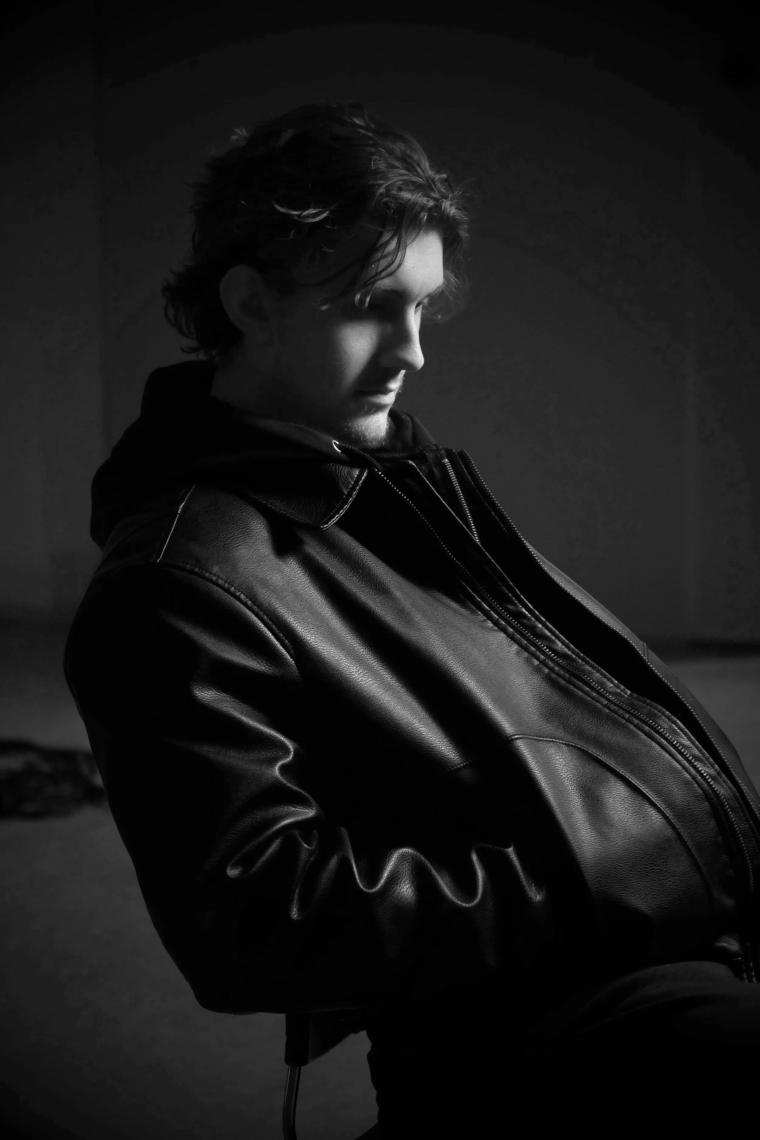 Black and white photo of a young man with curly hair wearing a leather jacket and hoodie, looking downward with a contemplative expression.