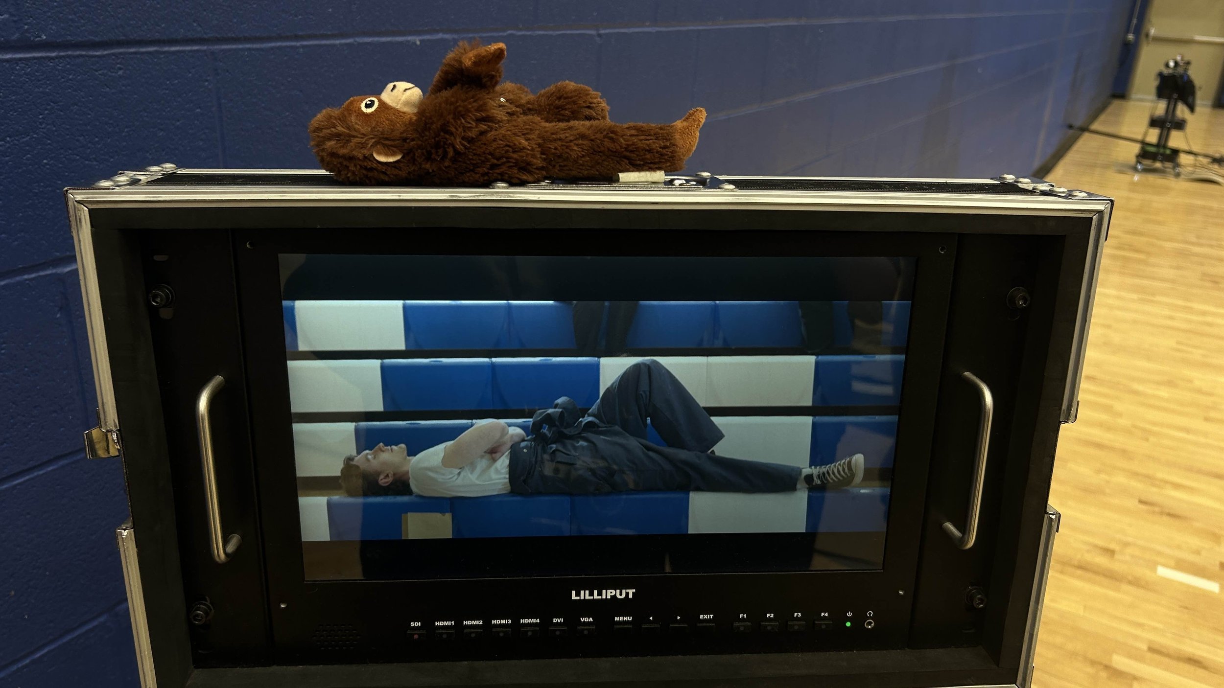 A child lying on a blue and white bench inside a glass enclosure in a gymnasium, with a stuffed bear toy on top of a black case nearby.