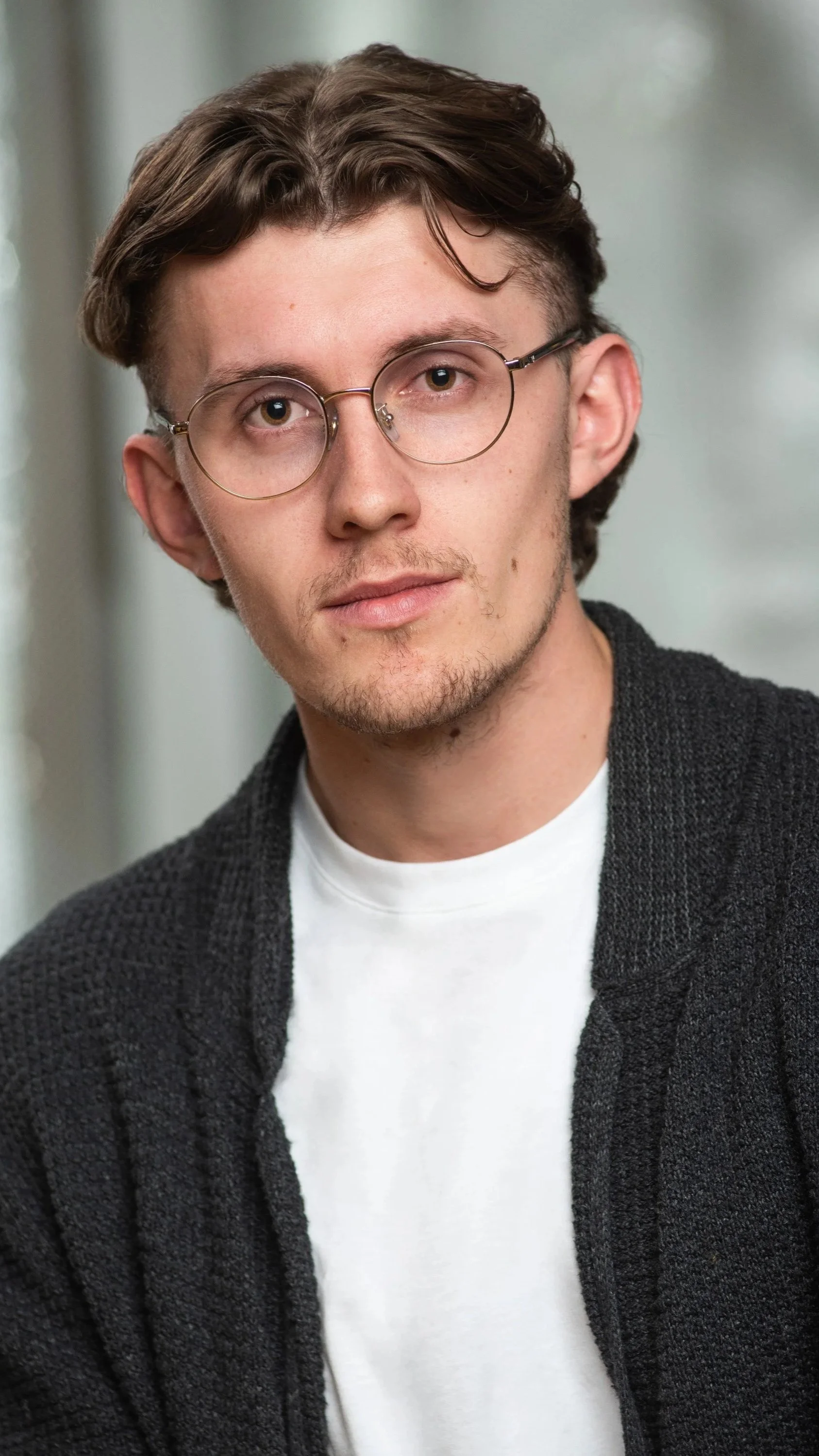 A young man with light brown, wavy hair wearing round glasses, a white shirt, and a dark knitted cardigan.