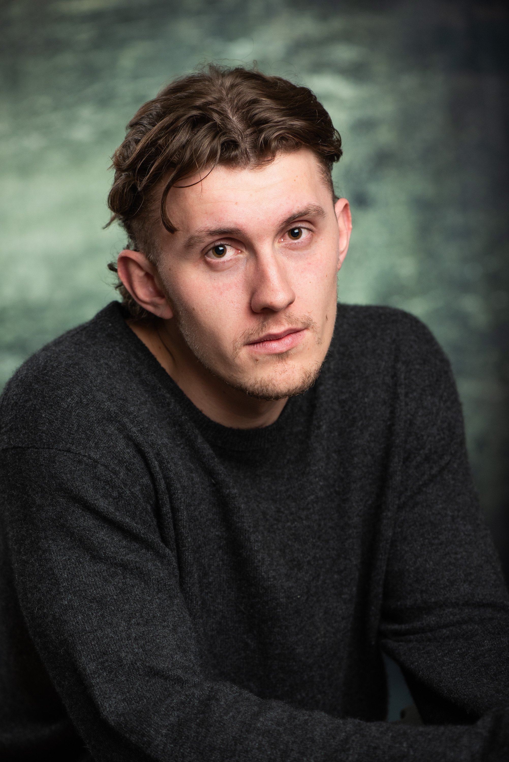 Portrait of a young man with curly brown hair, wearing a dark sweater, looking directly at the camera against a blurred green background.