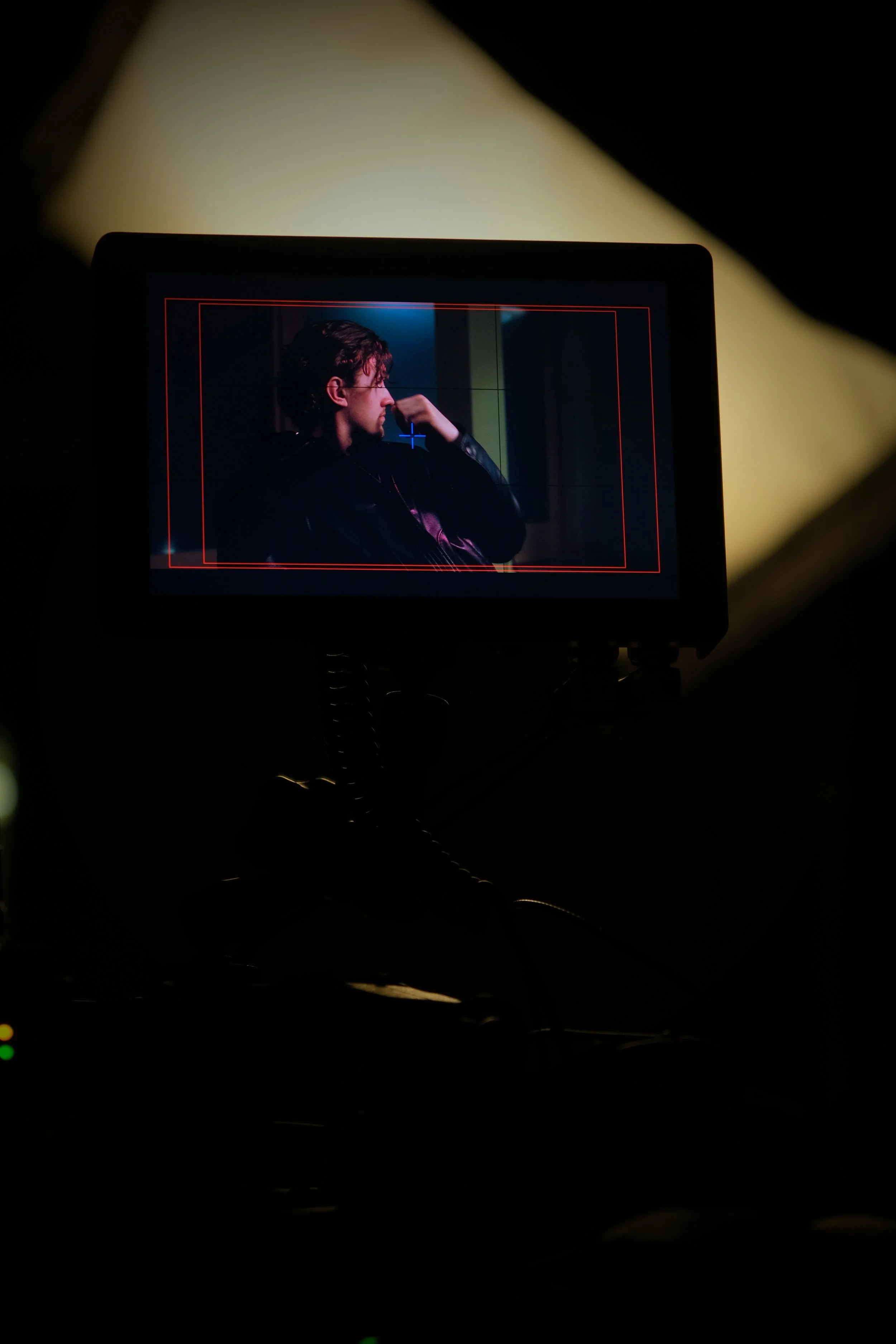 A person on a film or video shoot, seen through a camera monitor, sitting and thinking with a hand near their face, with a dark background and moody lighting.