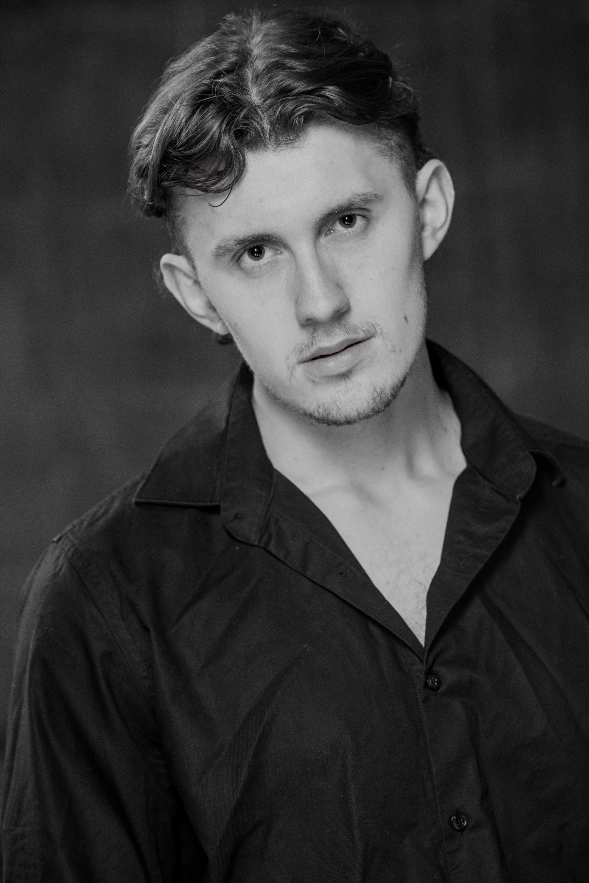 Black and white portrait of a young man with curly hair, wearing a dark shirt, looking into the camera with a serious expression.
