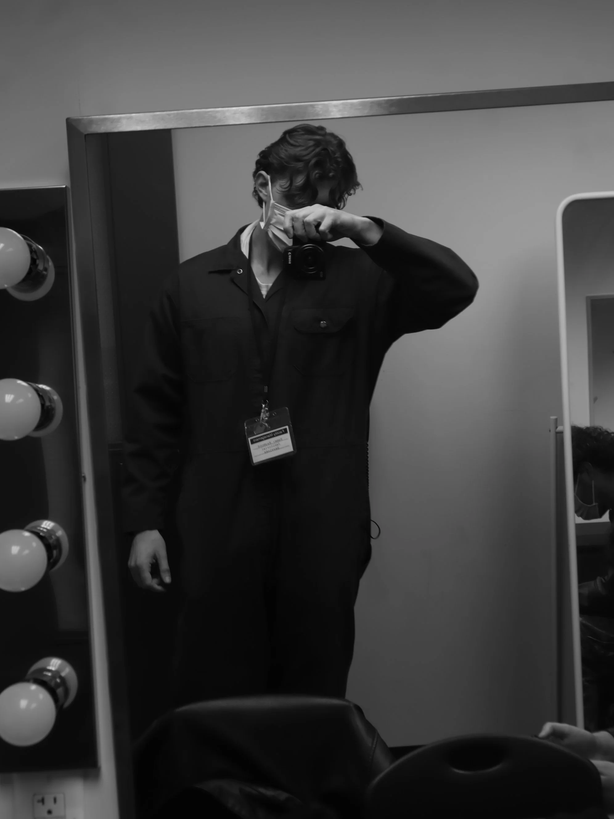 A person taking a selfie in a mirror, wearing a face mask, black jacket, and identification badge, with salon or dressing room lights visible on the side.