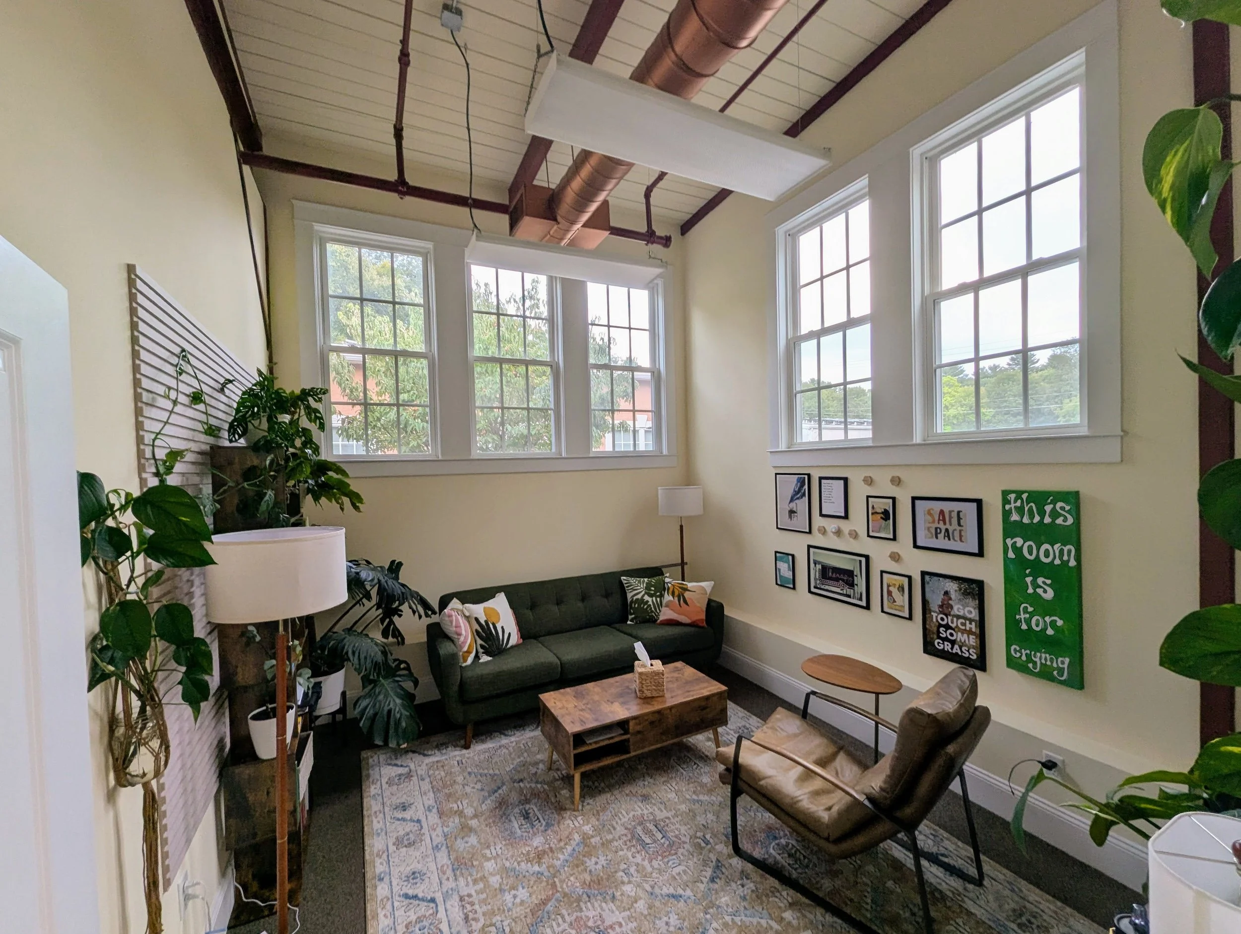 A cozy living room with large windows, a dark green sofa decorated with colorful cushions, a wooden coffee table, a leather armchair, potted plants, a gallery wall with framed pictures, and a sign that reads "This room is for crying".