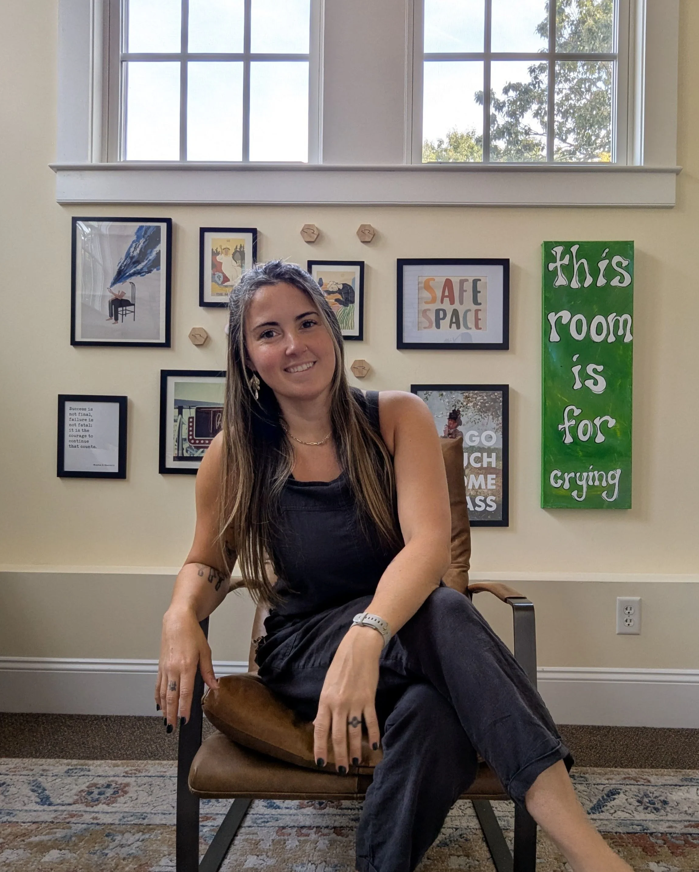 A smiling woman with long brown hair sitting on a brown chair in a room with a wall decorated with framed artwork and a green sign that reads "this room is for crying". There are two large windows above the wall, and she is wearing a black sleeveless top and dark jeans.