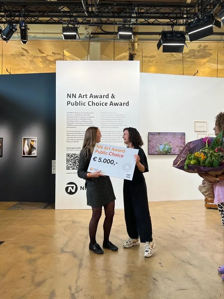 Two women, Inez de Brauw and Elisah van den Bergh, standing in an art gallery space, holding a large cheque for 5000 euros, with a woman holding a bouquet of flowers nearby. The background displays text about art awards and some artwork on the walls.