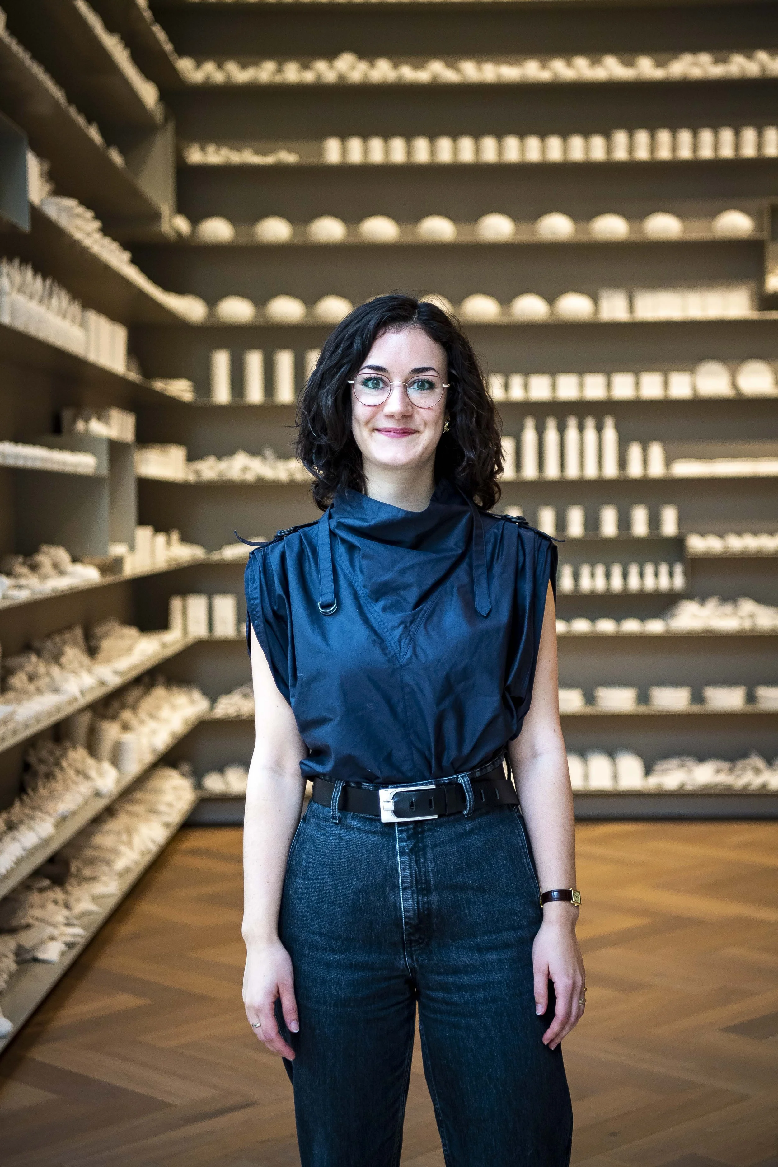 A smiling woman with glasses, dark curly hair, wearing a blue sleeveless top and high-waisted jeans, standing in front of shelves filled with white ceramic objects in a room with wooden flooring.