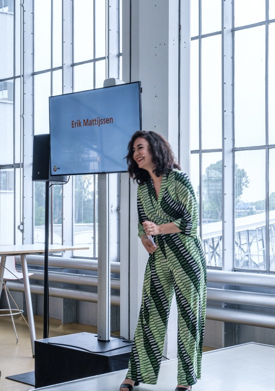 A woman, Elisah van den Bergh, laughing and standing next to a large screen displaying the name 'Erik Mattijssen' in a modern, glass-windowed indoor space.