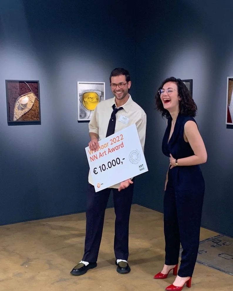 Two people standing in an art space, one man, Vytautas Kumza, holding a large cheque for 10,000 euros for the winning of the 2022 NN Art Award, both are smiling and laughing, with artworks displayed on the walls behind them.