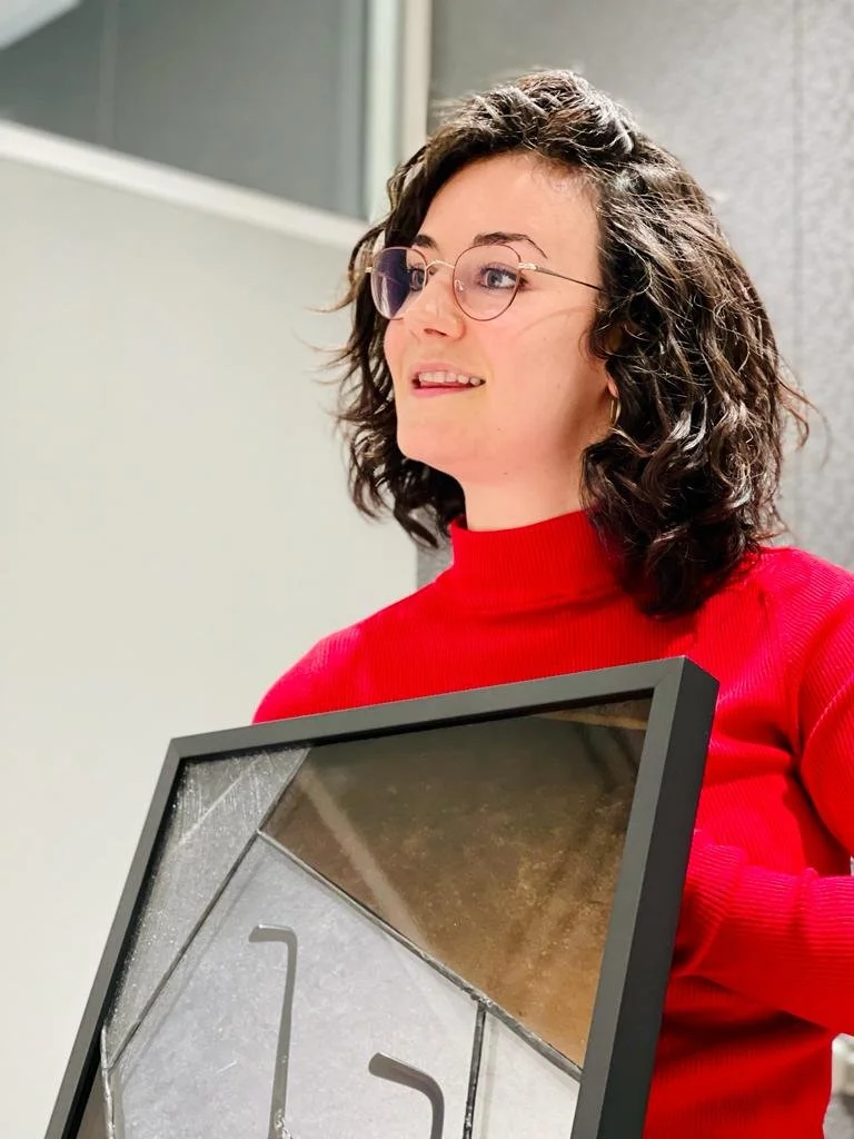 A woman with curly dark hair, glasses, and a red turtleneck sweater holding a frame or artwork by Vytautas Kumza, smiling in an indoor setting.