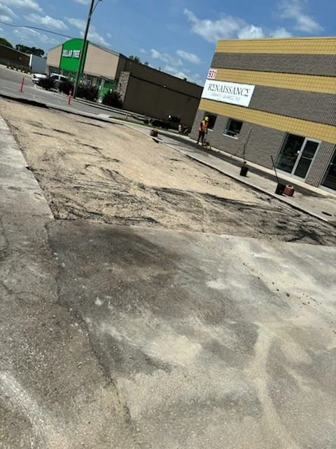 Street with a section of construction or pavement work, partially dug up ground, and two buildings in the background, one with signage for 'Rejuvenate' and another with a green sign.