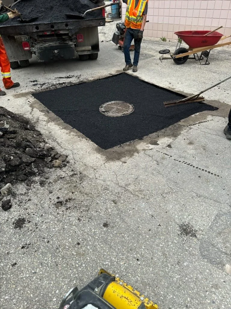 Workers pour fresh asphalt around a manhole cover on a street, with construction equipment and tools nearby.