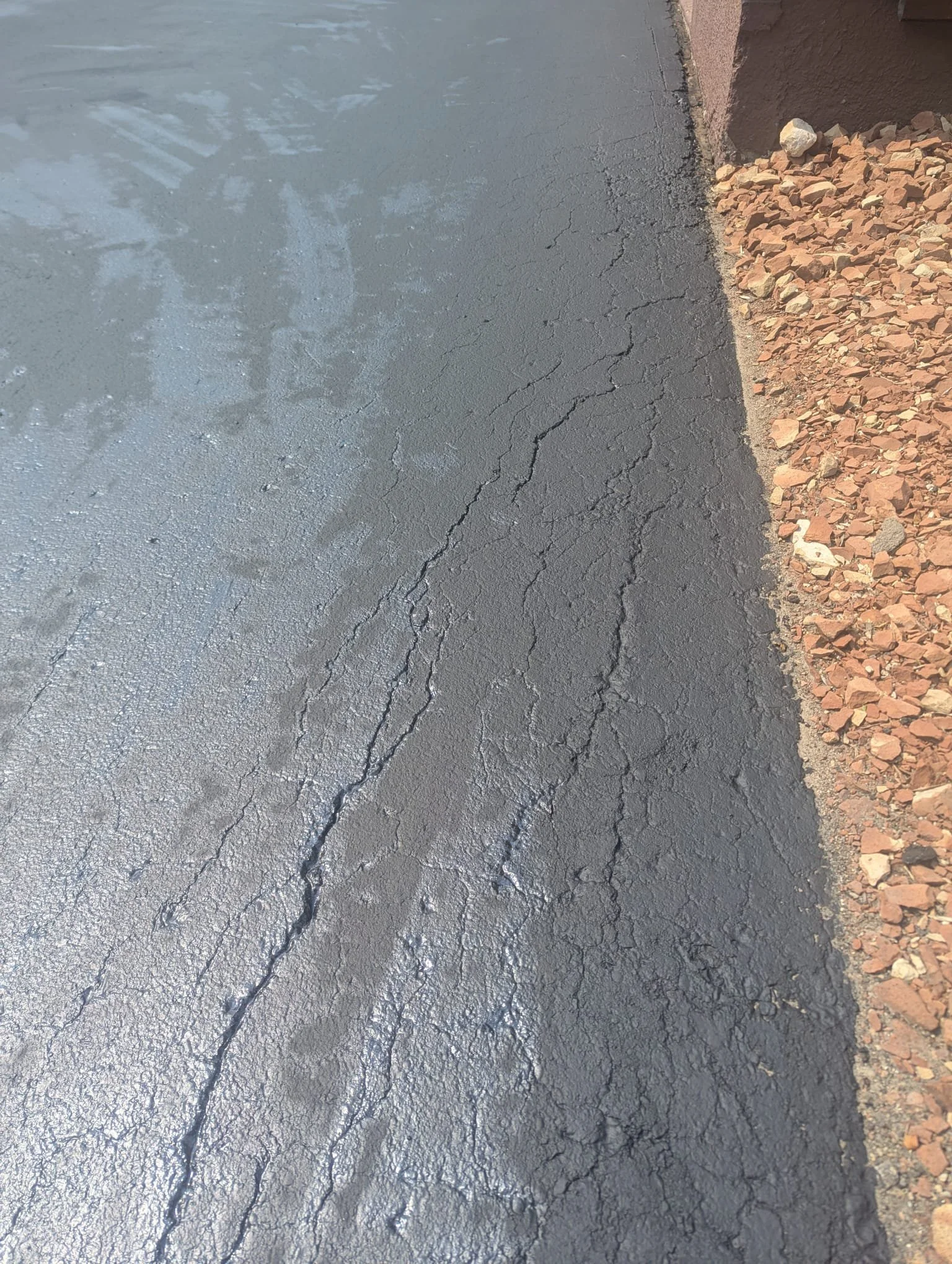 Close-up view of a freshly paved asphalt surface with visible cracks, next to a gravel area and a brick wall.