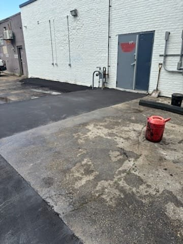 A partially paved area with a red bucket placed on the ground, next to a white brick building with a door and some pipes.