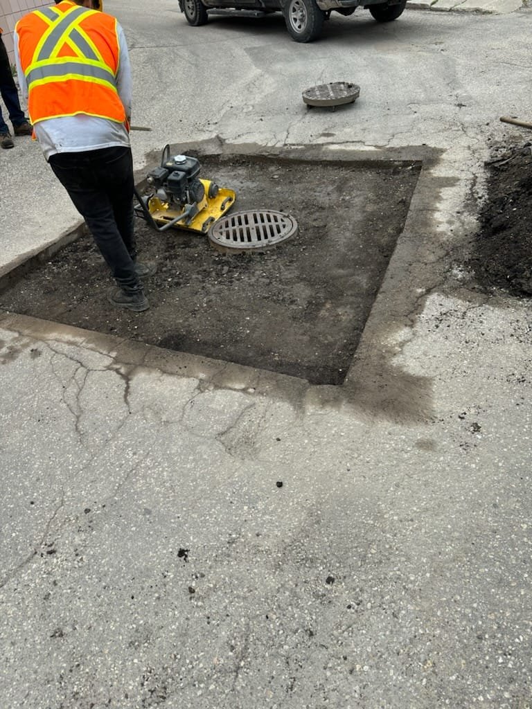 Worker in safety vest repairing a flooded storm drain on a street with a mini compactor and a manhole cover removed