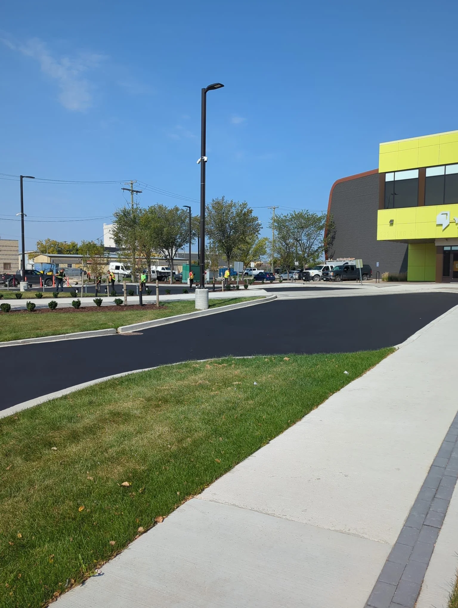 A newly paved black asphalt sidewalk next to a grassy area with small bushes and young trees, with a modern building and parked cars in the background under a clear blue sky.