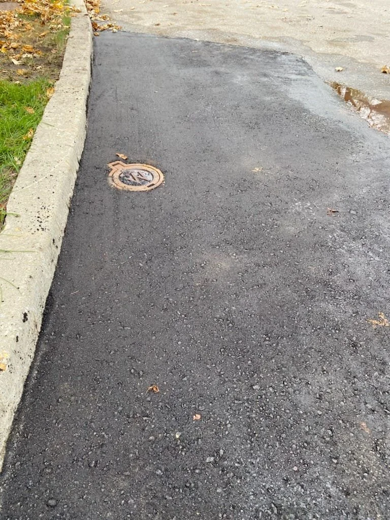 Freshly paved black asphalt sidewalk with a curb on the left side, grass with some fallen leaves, and a manhole cover near the curb.