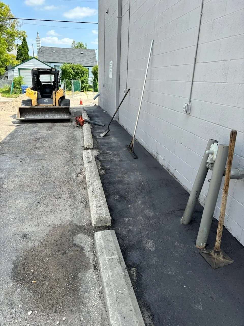 Construction site with a small yellow skid-steer loader, tools, and a freshly paved black asphalt strip along a gray building wall, with parking blocks and construction equipment.