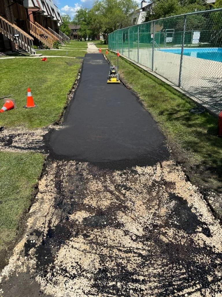 A newly paved sidewalk in an outdoor residential area, with construction cones and equipment visible, next to a grassy yard and a fenced swimming pool.