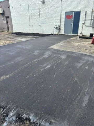 Freshly paved asphalt surface in an alley or parking lot area next to a white brick building with a blue door.