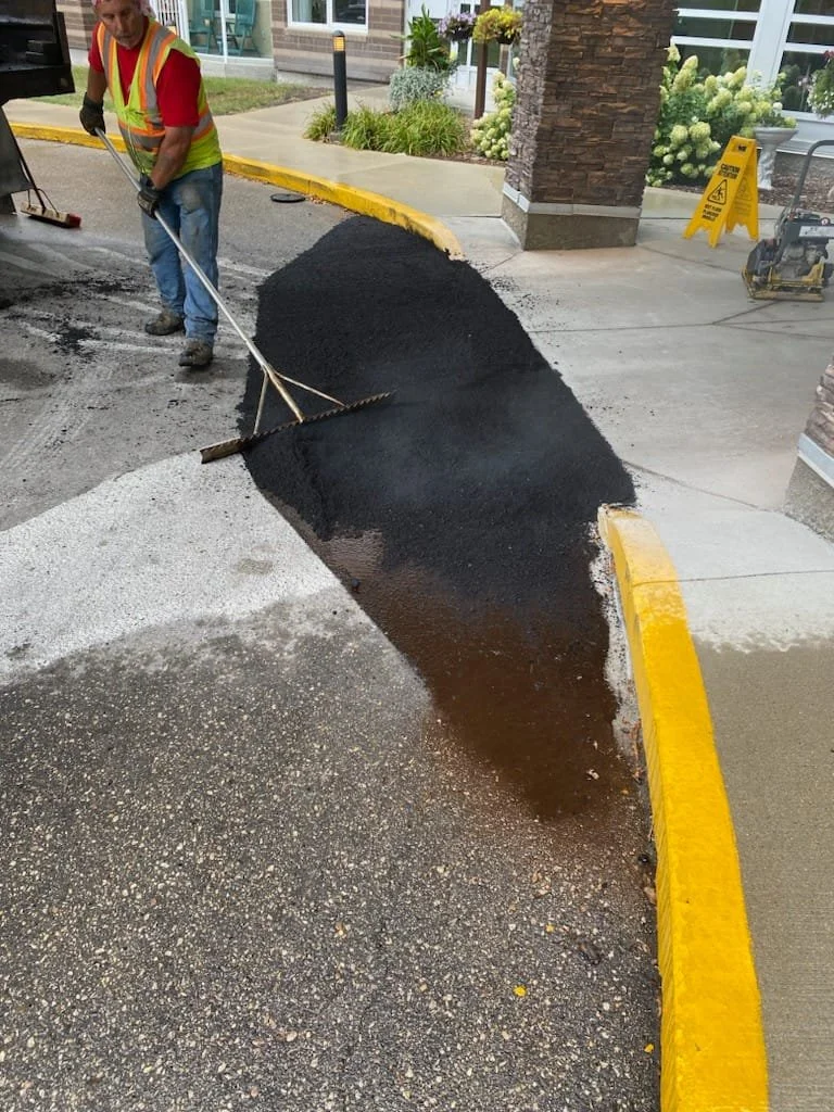 A construction worker in a yellow safety vest smoothing fresh black asphalt with a rake on a city street curb.