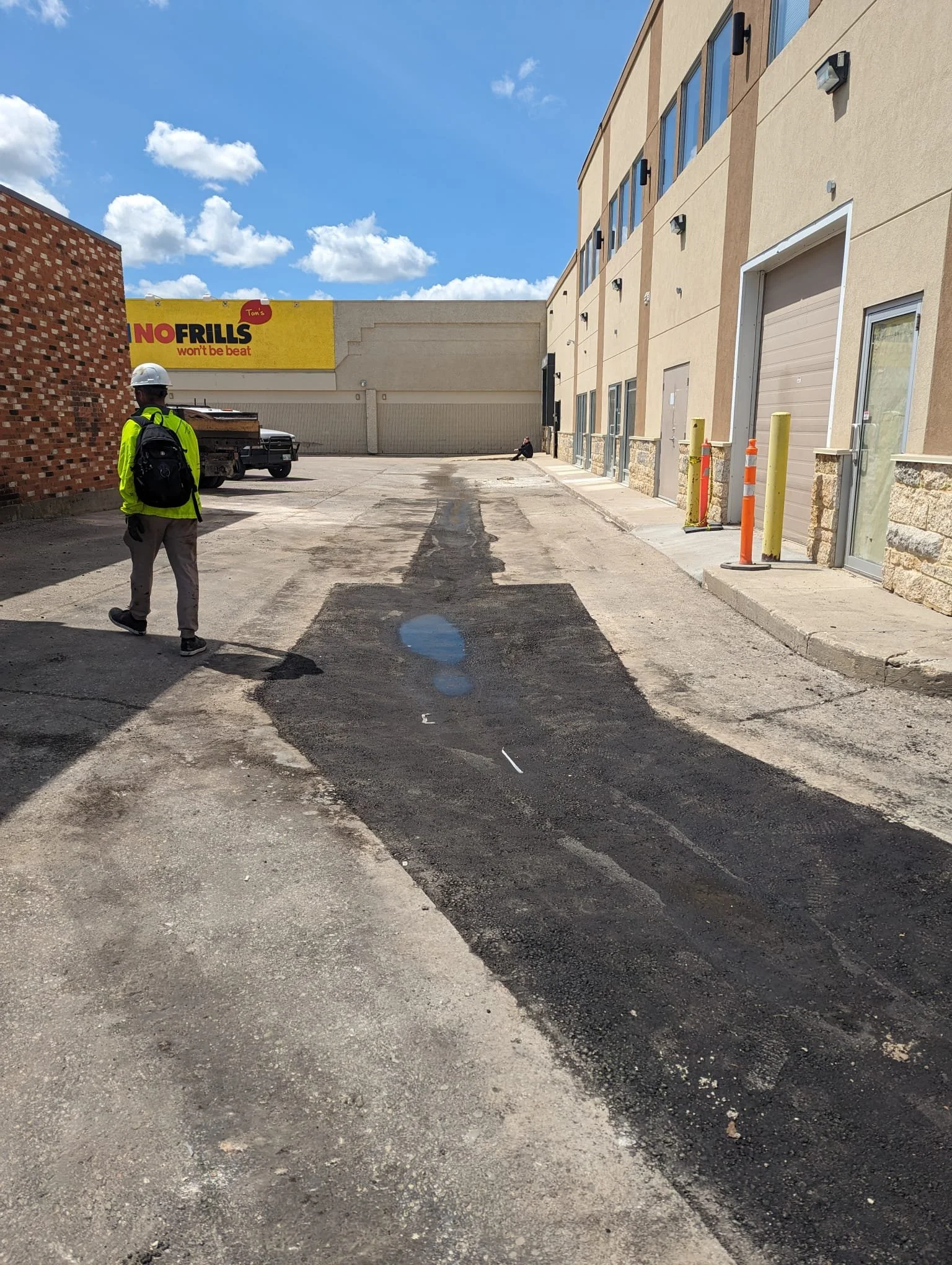 A man wearing a yellow safety jacket, gray pants, and a white helmet stands in a parking lot. There are puddles on the ground and construction equipment nearby. A person is sitting on the curb in the background, and a building with large windows and 