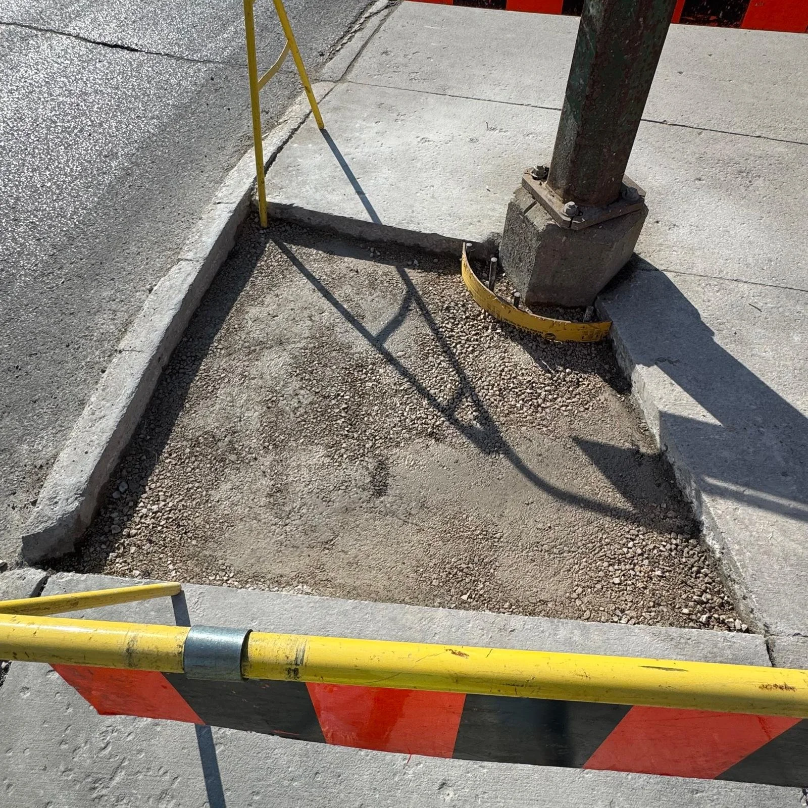 A parking lot with a concrete curb, a metal pole, and a yellow safety barrier. The ground has some gravel and concrete, and a shadow of the safety barrier is cast on the ground.