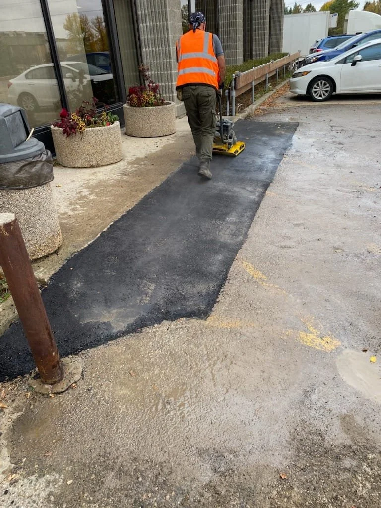 Worker in an orange safety vest operating a roller to asphalt a sidewalk outside a building with large glass windows and potted plants.