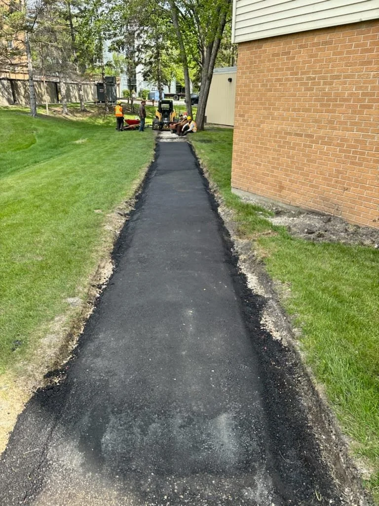 A newly paved asphalt sidewalk next to a brick building, with green grass on both sides, and construction workers in the background working on finishing the sidewalk.