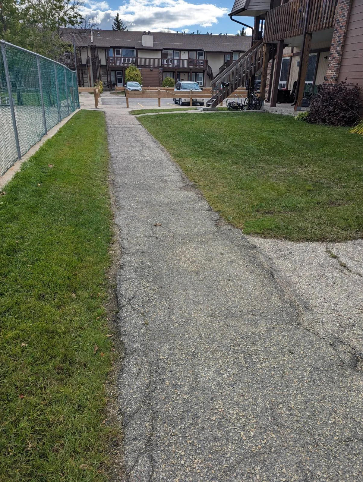 A cracked asphalt pathway leading to a parking lot and residential buildings in the background with parked cars and bicycles.