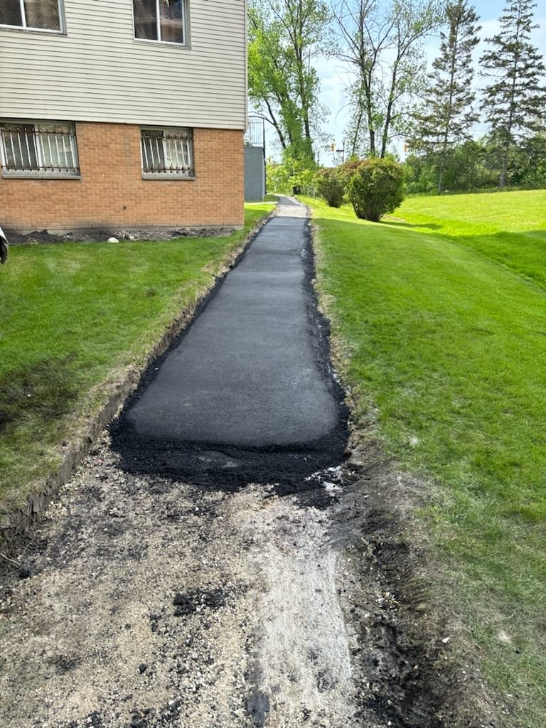 Newly paved sidewalk leading away from a house with green grass on both sides and trees in the background.