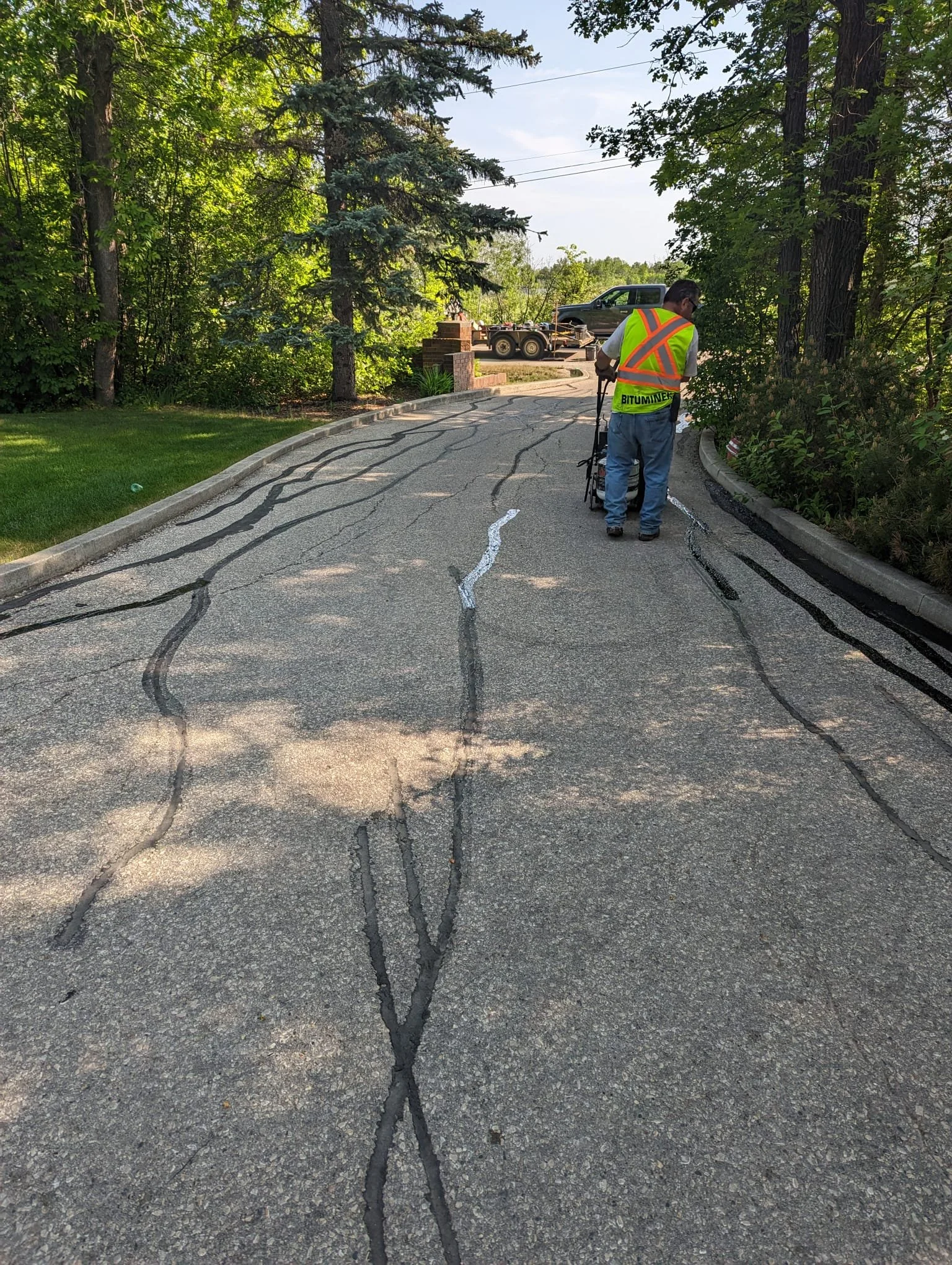 A worker in a yellow safety vest coloring on a cracked asphalt road, surrounded by trees.
