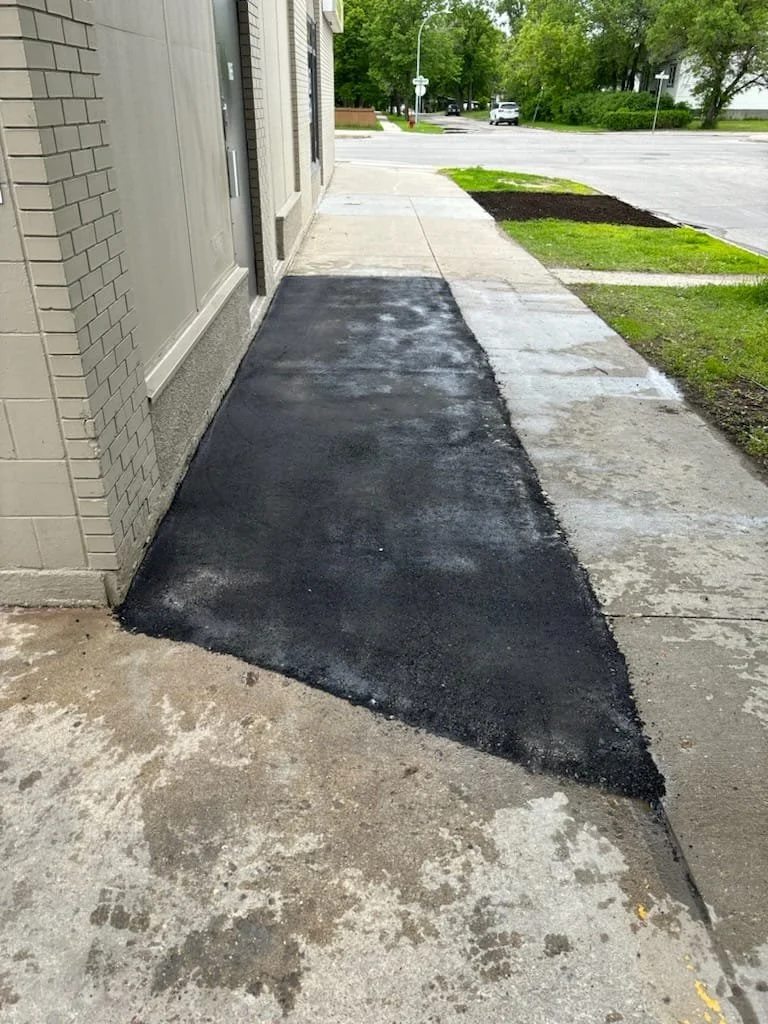 Freshly paved black asphalt patch on sidewalk next to a beige brick building, with a street and parking lot in the background.
