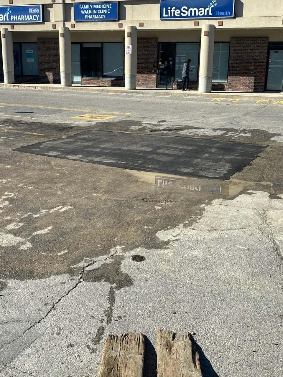 A small parking lot in front of a pharmacy with signs for family medicine, walk-in clinic, and pharmacy. The parking lot has a large patch of fresh asphalt and a small puddle. A person is walking near the pharmacy entrance.