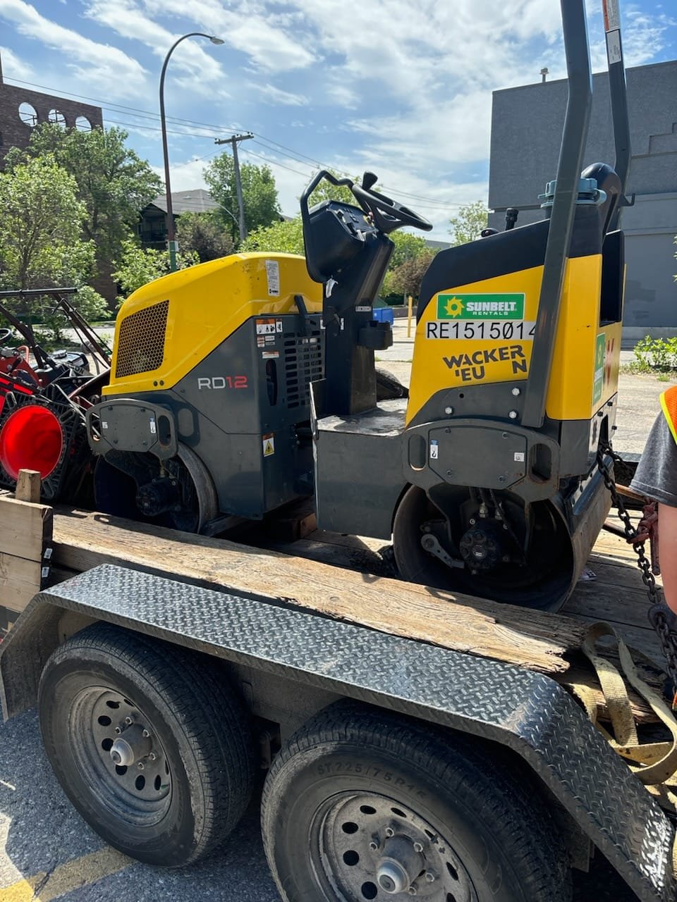 A small yellow and black Wacker RD12 ride-on roller compactor on a flatbed trailer in a parking lot with green trees, a building, and a blue sky with clouds in the background.