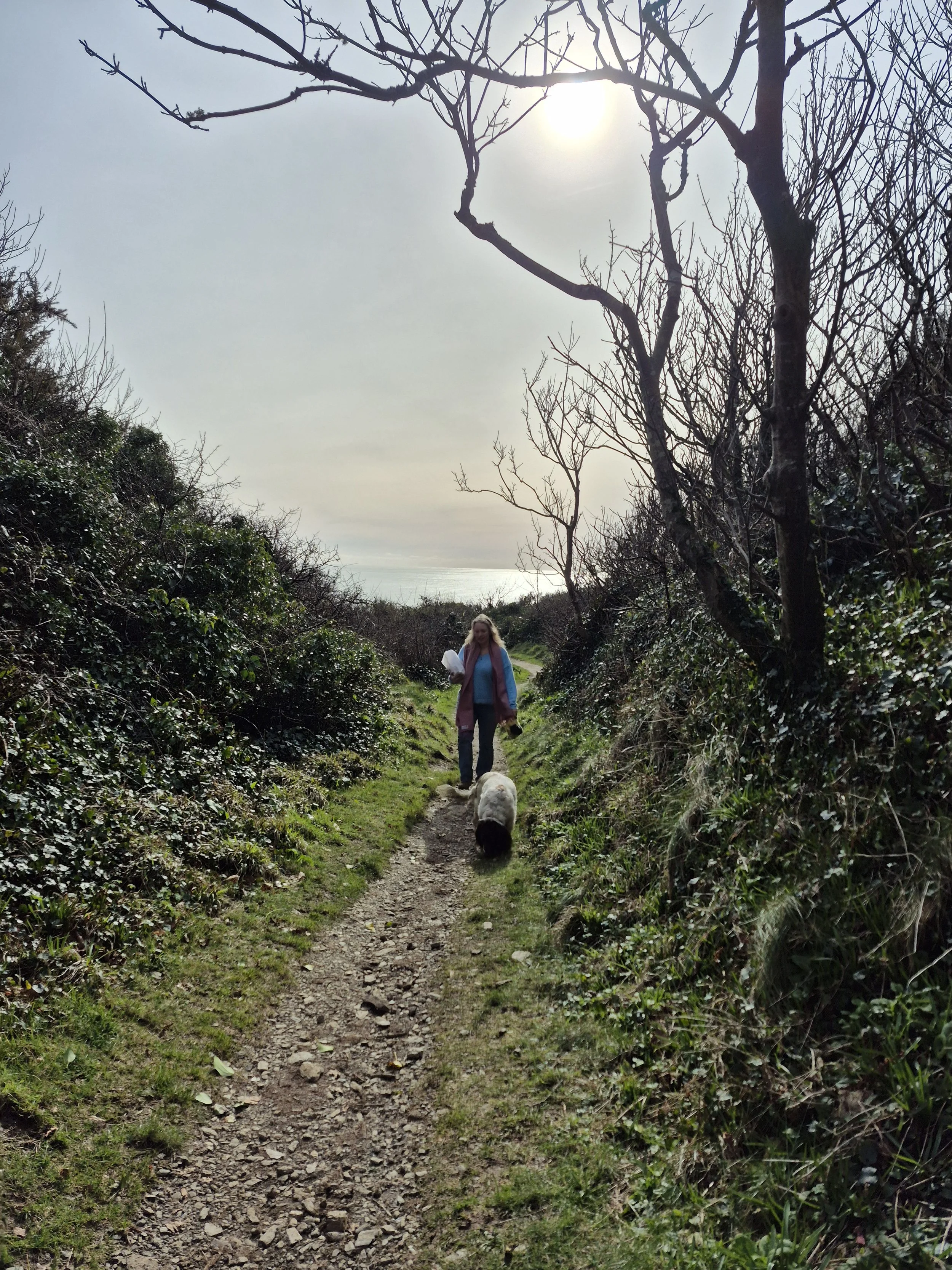 Laura Jessica walker walking her dog along a dirt trail surrounded by bushes and leafless trees, with the sun shining through the sky in a cloudy day. Sea behind her.
