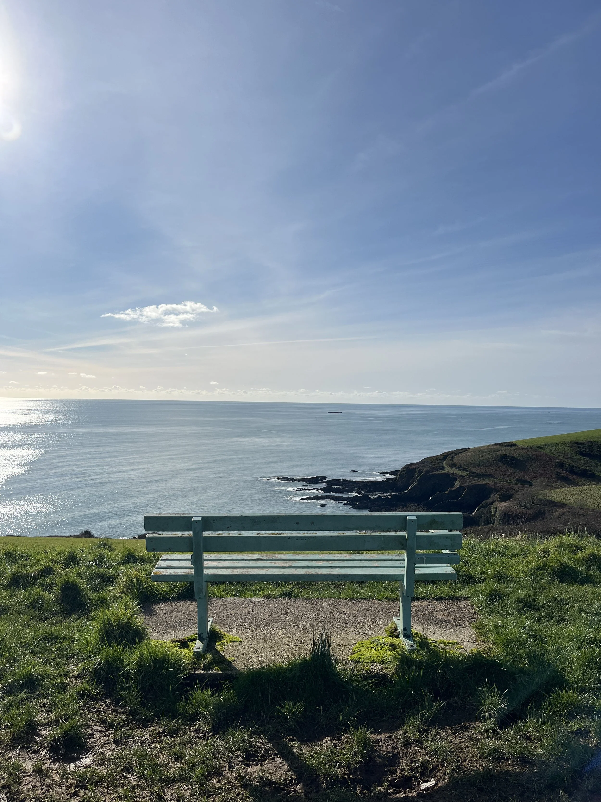 A turquoise bench facing the Cornish ocean on a grassy hill with cliffs, under a partly cloudy blue sky.